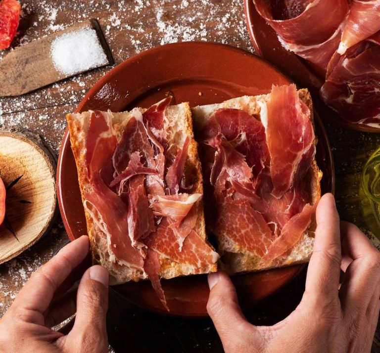 high angle view of a man preparing a typical spanish bocadillo de jamon, a serrano ham sandwich, on a rustic wooden table, next to a plate with some slices of serrano ham and a cruet with olive oil (high angle view of a man preparing a typical spanish